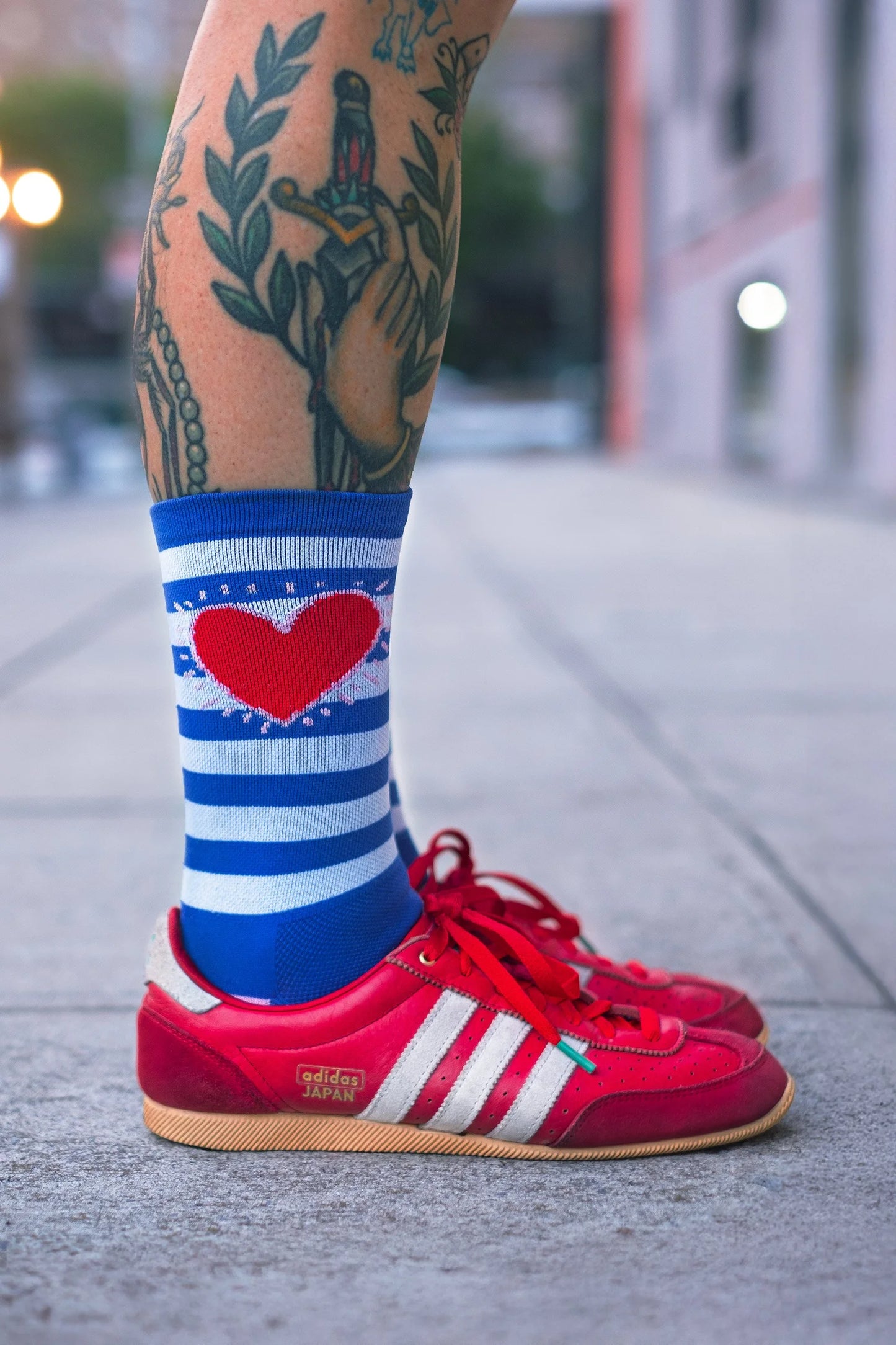 Person wearing red Adidas sneakers with side view of Affinity x Keith Haring “Stripe Heart” blue and white striped cycling socks with red heart, worn by tattooed cyclist, on a blurred street background.