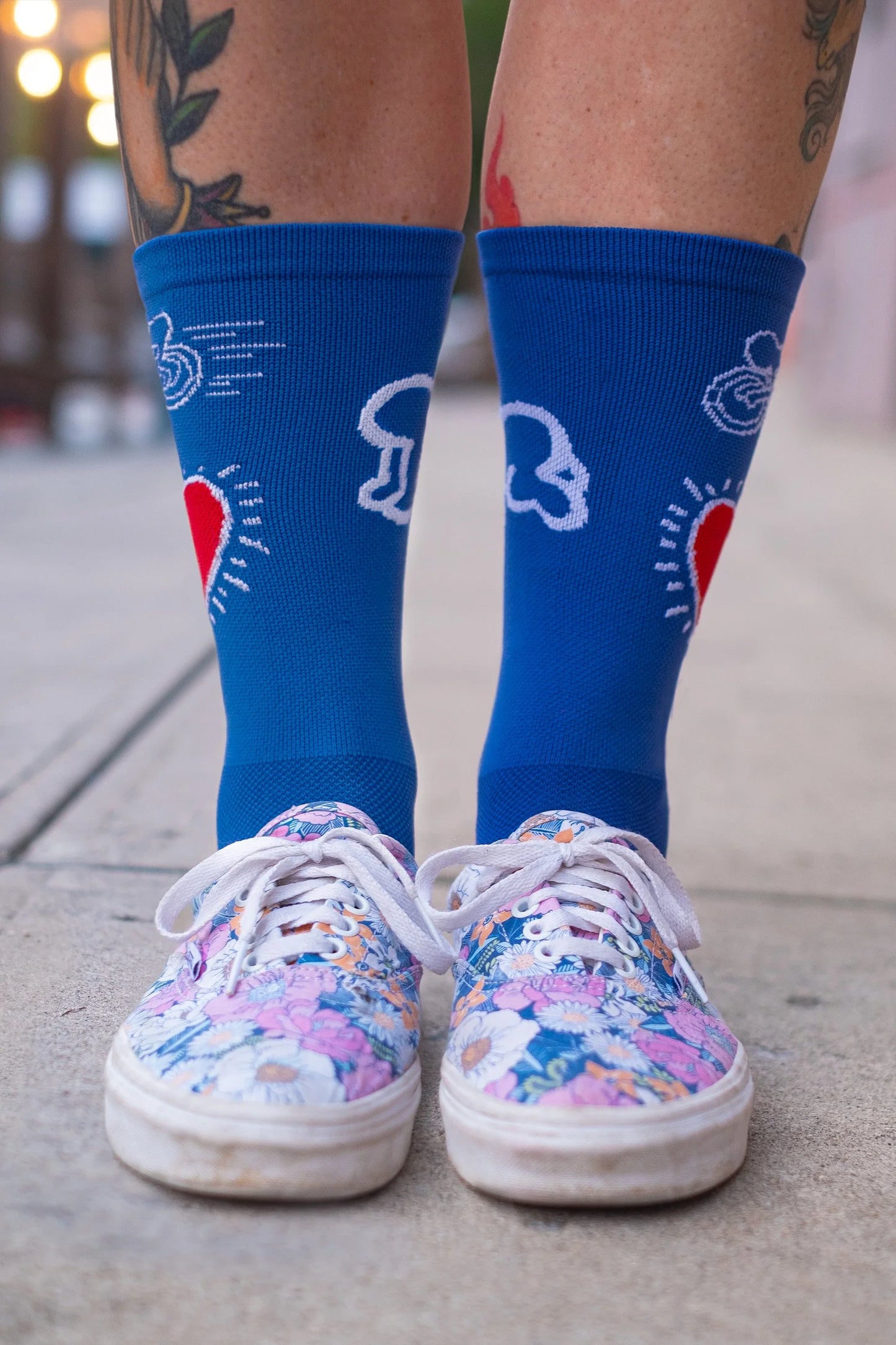 Front close-up of Affinity x Keith Haring “Bike Heart” royal blue cycling socks with red heart and bike graphic, worn with floral sneakers.