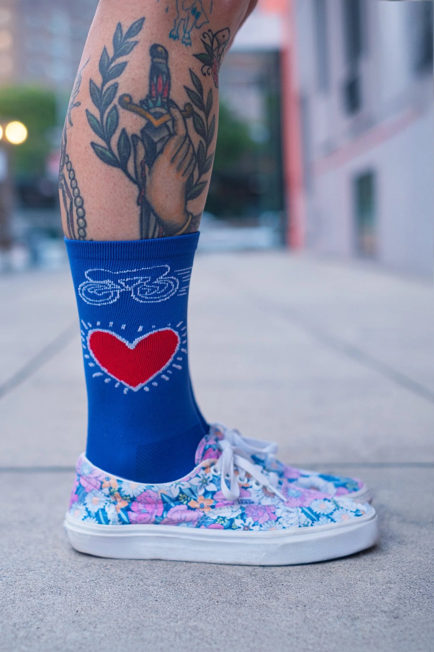 Close-up of Affinity x Keith Haring “Bike Heart” royal blue cycling socks with red heart and bike graphic, worn with floral sneakers.