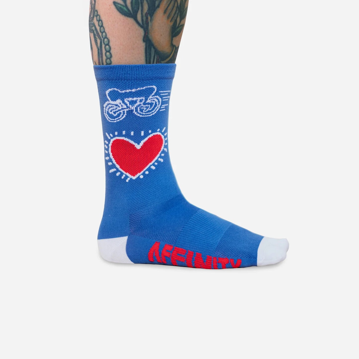 Close-up of Affinity x Keith Haring “Bike Heart” royal blue cycling socks with red heart and bike graphic, worn with floral sneakers.