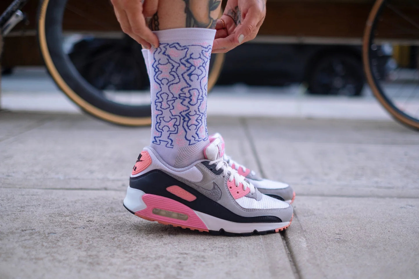 Side view of cyclist wearing Affinity x Keith Haring “Dancing Figures” white and pink cycling socks with Haring’s iconic outlined characters, paired with pink Nike Air Max shoes.