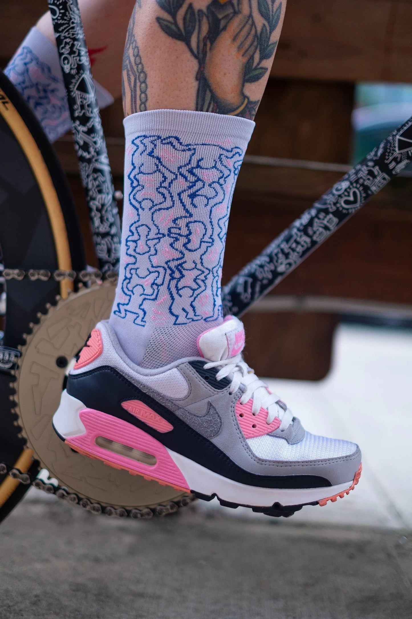 Cyclist wearing Affinity x Keith Haring “Dancing Figures” white and pink cycling socks with Haring’s iconic outlined characters, paired with pink Nike Air Max shoes.