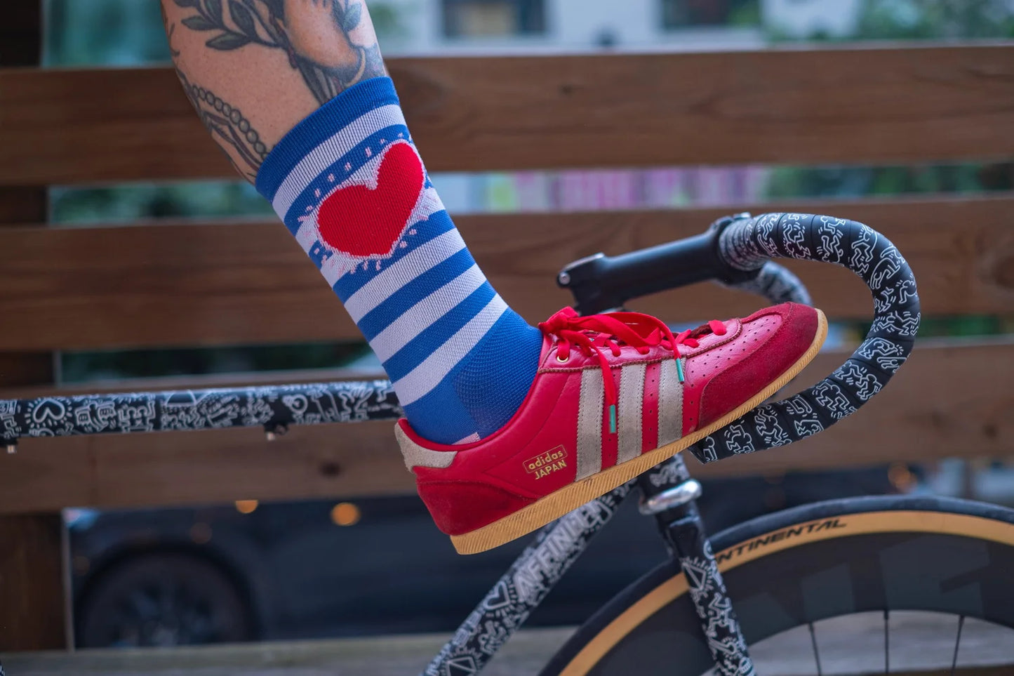 Side view of Affinity x Keith Haring “Stripe Heart” blue and white striped cycling socks with red heart, worn by tattooed cyclist, wearing red Adidas sneakers.