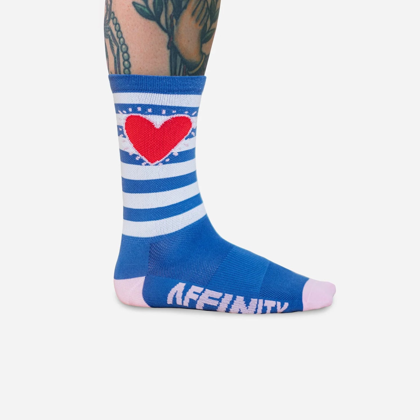 Side view of Affinity x Keith Haring “Stripe Heart” blue and white striped cycling socks with red heart, worn by tattooed cyclist, on solid white background.