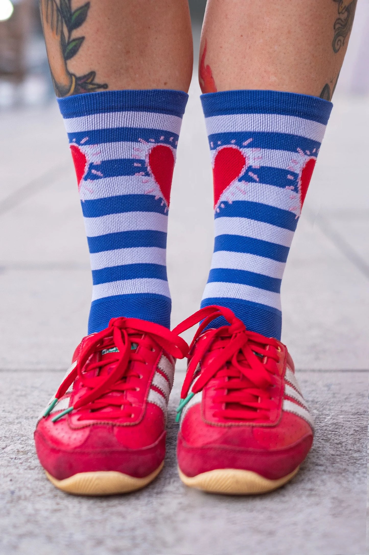 Front view of Affinity x Keith Haring “Stripe Heart” blue and white striped cycling socks with red heart, worn by tattooed cyclist. On a light background.