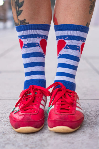 Front view of Affinity x Keith Haring “Stripe Heart” blue and white striped cycling socks with red heart, worn by tattooed cyclist. On a light background.