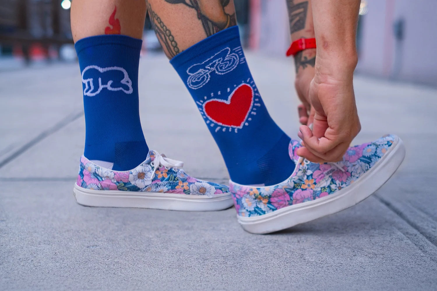 Close-up of Affinity x Keith Haring “Bike Heart” royal blue cycling socks with red heart and bike graphic, worn with floral sneakers.
