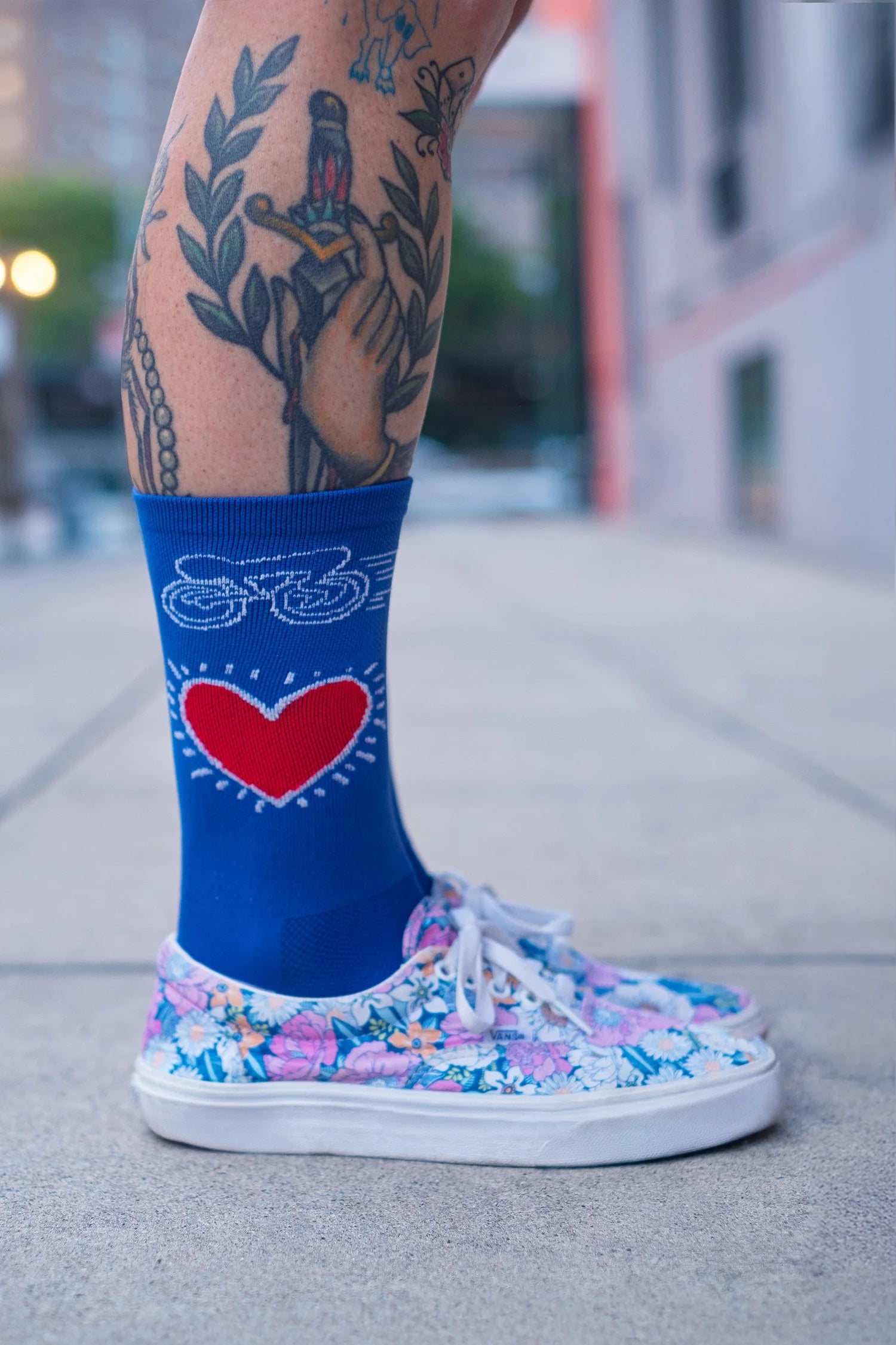 Close-up of Affinity x Keith Haring “Bike Heart” royal blue cycling socks with red heart and bike graphic, worn with floral sneakers.