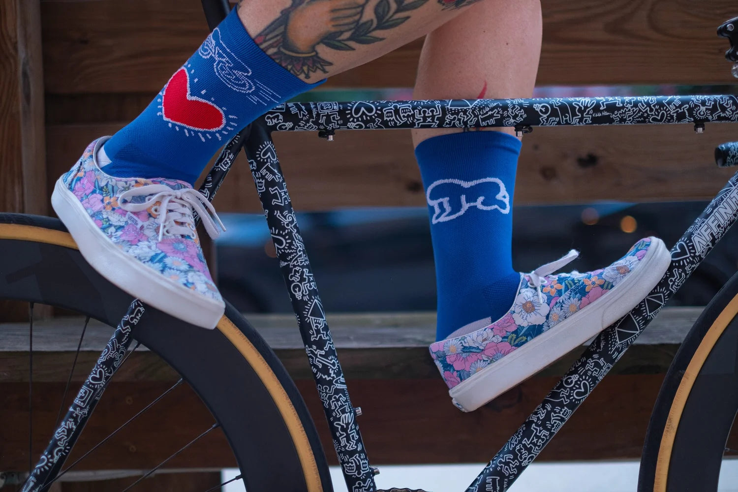 Close-up of Affinity x Keith Haring “Bike Heart” royal blue cycling socks with red heart and bike graphic, worn with floral sneakers.