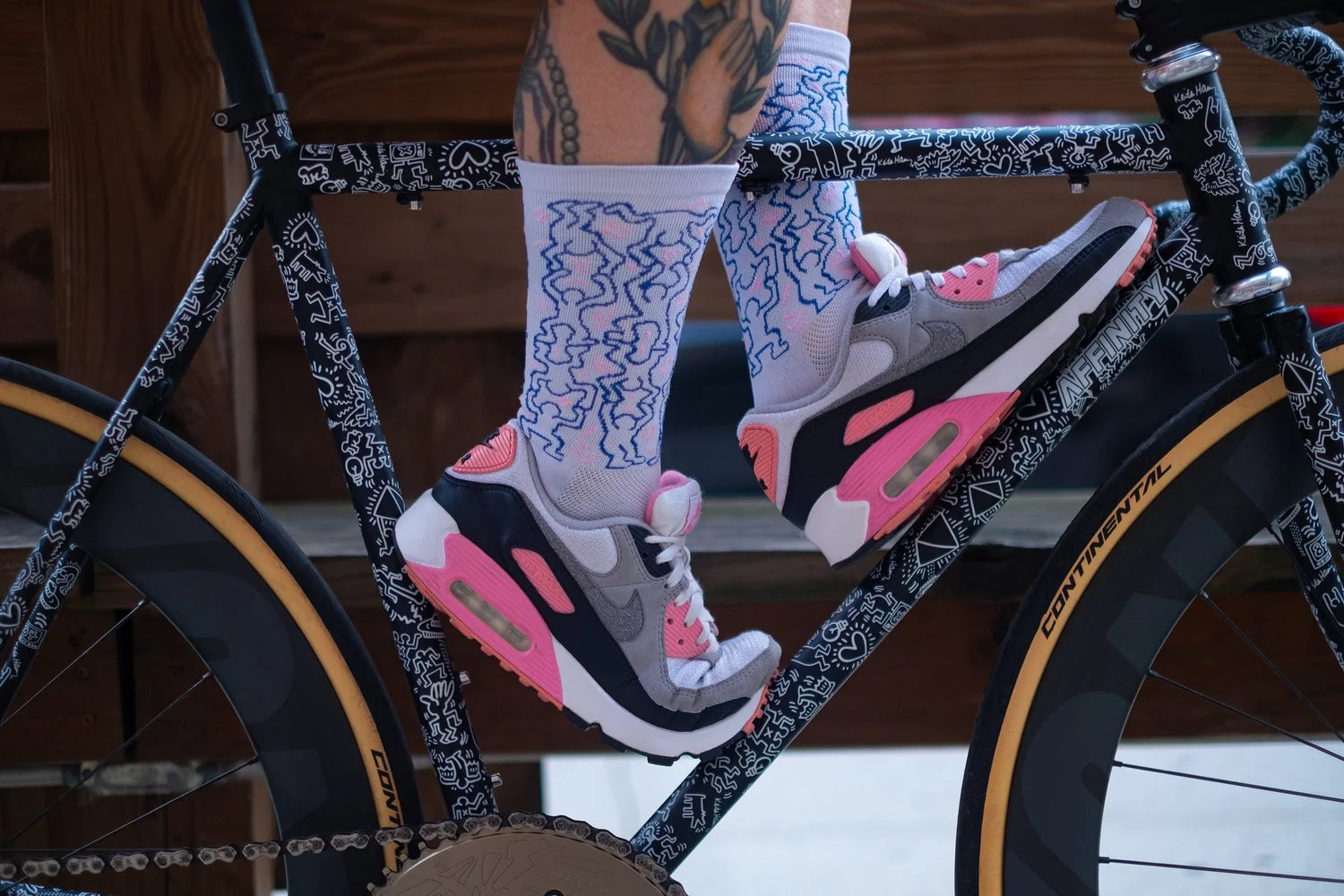 Cyclist wearing Affinity x Keith Haring “Dancing Figures” white and pink cycling socks with Haring’s iconic outlined characters, paired with pink Nike Air Max shoes.